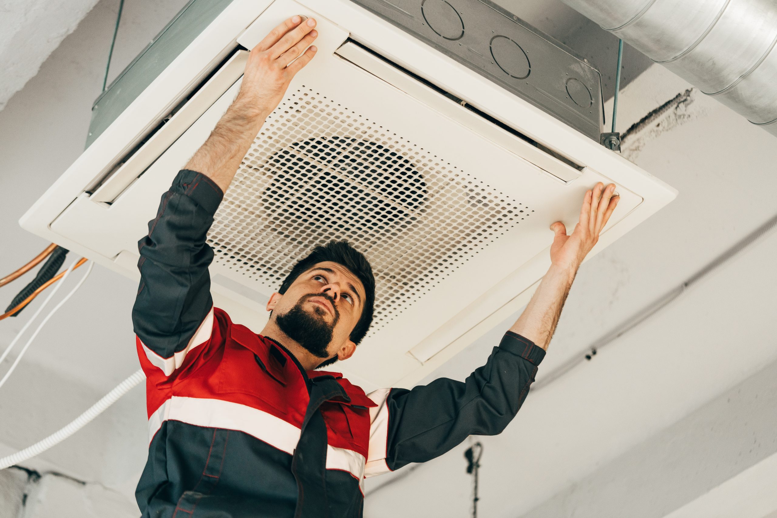 A technician is focused on installing a ceiling-mounted air conditioning unit in a contemporary office setup. The workspace is well-lit and features exposed ductwork.